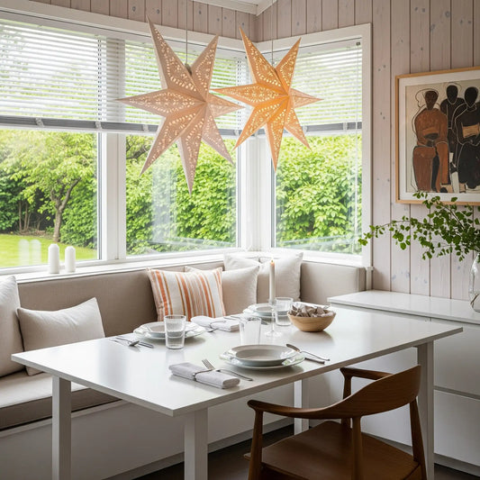 Sunlit breakfast nook with a built-in bench, white table set for two, and a wooden chair. Two beige paper star lanterns hang above. Large windows show a green garden. A framed painting and a sprig of greenery add subtle detail.