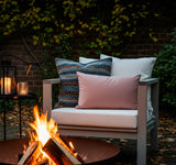 Cozy outdoor evening scene with a burning fire pit in the foreground. Behind it, a light wood armchair with white cushions and blue, pink, and white pillows. A black side table with lanterns sits to the left. -Square Custom Seat Cushions-Custom Rectangle Seat Cushion-Pet-Resistant Dining Cushion
