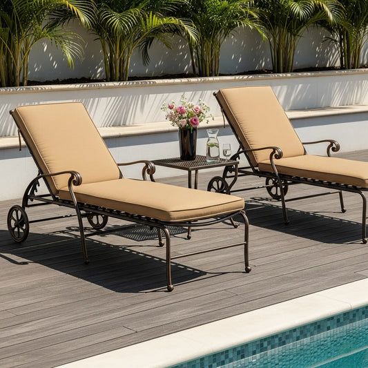 Two black wrought iron chaise lounges with tan cushions sit on a dark wood deck beside a pool. A matching side table between them holds a pitcher and a vase of flowers. A white wall with lush green palm plants is in the background.