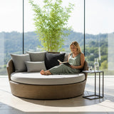 A blonde woman in a green dress reads a book while relaxing on a large, round, woven daybed with light grey cushions. The daybed is in a sunlit room with a floor-to-ceiling window showing a green landscape. A small side table holds a glass of water.