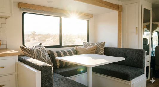 The bench cushions are upholstered in durable, textured charcoal grey fabric, accented with stylish patterned throw pillows. Sunlight streams through the large RV window creating a warm, inviting atmosphere. High resolution, interior design photography.