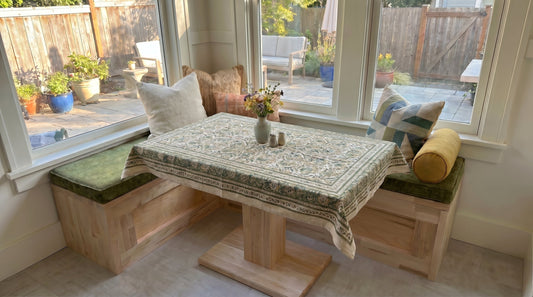 Breakfast nook bench cushion on a custom built-in corner seating area with green seat cushions, decorative pillows, and a wooden dining table by large windows