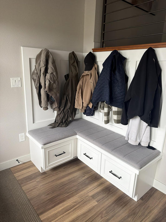 L-shaped built-in mudroom bench with gray custom cushion, coats on hooks above, storage drawers below