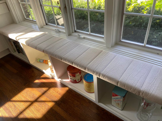 Single long custom window seat cushion wrapping a corner bench over open storage cubbies