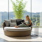 A blonde woman in a green dress reads a book while relaxing on a large, round, woven daybed with light grey cushions. The daybed is in a sunlit room with a floor-to-ceiling window showing a green landscape. A small side table holds a glass of water.
