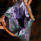 Side view of purple metallic handbag showing dimensional gathered fabric design